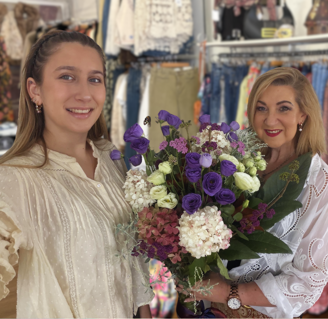 Lisa and India smiling in a clothing boutique, with the woman on the right holding a large bouquet of purple, white, and pink flowers. Clothing racks and mannequins are visible in the background.